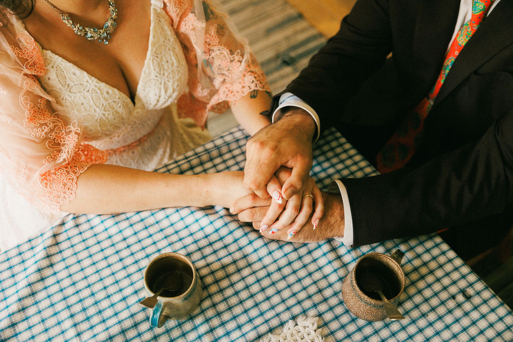 A bride and groom hold hands while sitting at a table with coffee mugs.